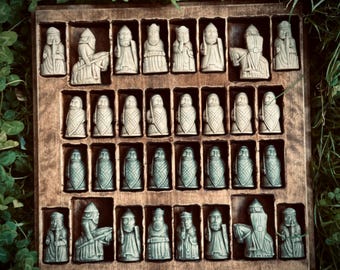 Puede incluir: Un juego de ajedrez completo con piezas de piedra tallada en una caja de madera. Las piezas son en tonos grises y verdes, que representan figuras con ropa y rasgos faciales detallados. El juego incluye un rey, una reina y otras piezas de ajedrez.