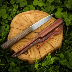 May include: A fixed-blade knife with a straight, silver-coloured metal blade and a dark brown wooden handle, accompanied by a matching brown leather sheath featuring visible stitching and a thin leather cord tie, resting on a round wood slice.
