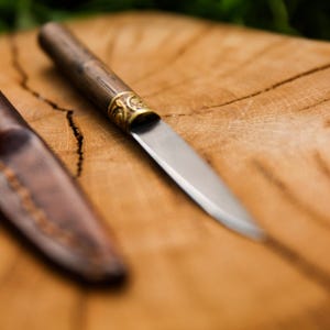 Puede incluir: Un cuchillo peque&ntilde;o de hoja fija con mango de madera marr&oacute;n oscuro, una virola de metal dorado con un patr&oacute;n de volutas en relieve y una hoja de acero afilada, acompa&ntilde;ado de una funda de cuero marr&oacute;n a juego con costuras visibles.