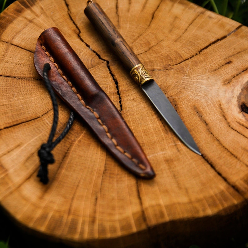 Puede incluir: Un cuchillo peque&ntilde;o de hoja fija con mango de madera oscura, virola de lat&oacute;n con patr&oacute;n de hojas en relieve y hoja de acero puntiaguda. Junto al cuchillo hay una funda de cuero marr&oacute;n con costuras visibles y un cord&oacute;n negro.