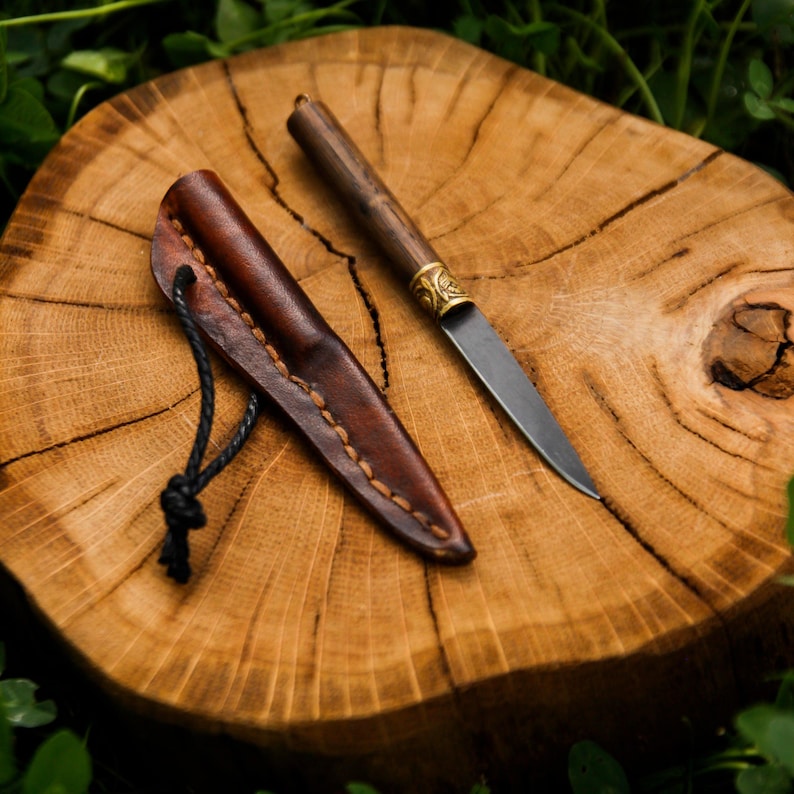 Puede incluir: Un cuchillo peque&ntilde;o de hoja fija con mango de madera oscura, virola de lat&oacute;n y hoja de acero puntiaguda. Junto al cuchillo hay una funda de cuero marr&oacute;n con costuras visibles y un lazo de cord&oacute;n negro para sujetarlo o transportarlo.