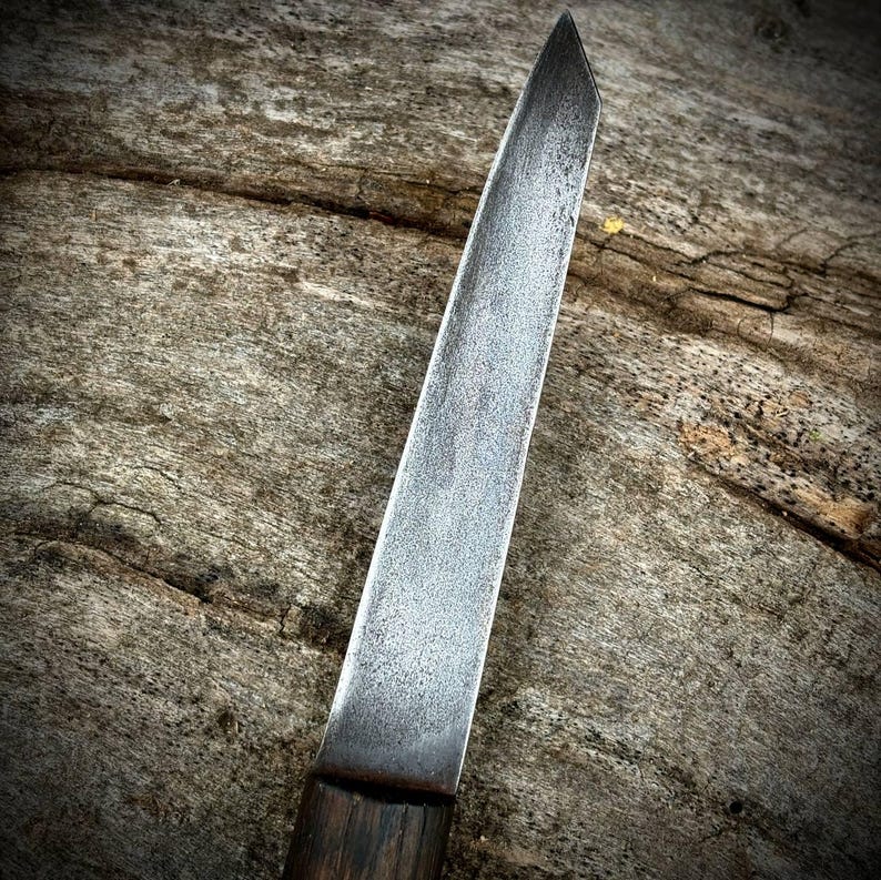 Puede incluir: Primer plano de un cuchillo largo, delgado y plateado con mango de madera. El cuchillo es afilado y tiene una hoja ligeramente curvada.