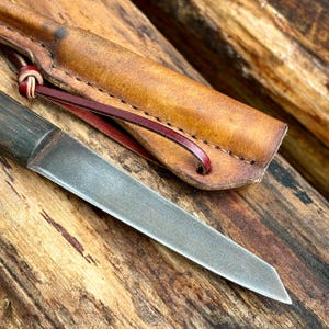 Puede incluir: Un cuchillo peque&ntilde;o, delgado, de plata con mango de madera y funda de cuero. El cuchillo est&aacute; sobre una superficie de madera.