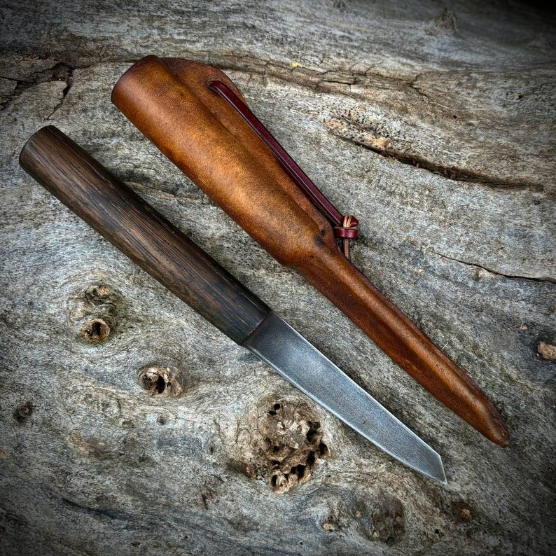 Puede incluir: Un peque&ntilde;o cuchillo artesanal con mango de madera oscura y una hoja fina y afilada. El cuchillo est&aacute; apoyado sobre un trozo de madera desgastada, y una funda de cuero marr&oacute;n con una correa roja est&aacute; a su lado.