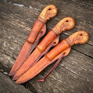 Op de afbeelding: Drie handgemaakte messen met houten handvatten en leren scheden. De handvatten hebben ingewikkelde gesneden ontwerpen en de scheden zijn een rijke bruine kleur. De messen worden gepresenteerd op een verweerd houten oppervlak, dat het vakmanschap laat zien.
