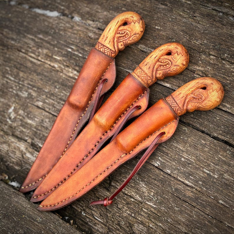 Op de afbeelding: Drie handgemaakte messen met houten handvatten en leren scheden. De handvatten hebben ingewikkelde gesneden ontwerpen en de scheden zijn een rijke bruine kleur. De messen worden gepresenteerd op een verweerd houten oppervlak, dat het vakmanschap laat zien.