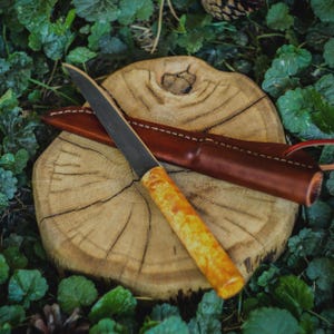 Puede incluir: Un cuchillo de hoja fija con una hoja de acero oscuro y un mango de madera texturizada de color claro, acompañado de una funda de cuero marrón con costuras visibles en los bordes, sobre una rodaja de madera circular.