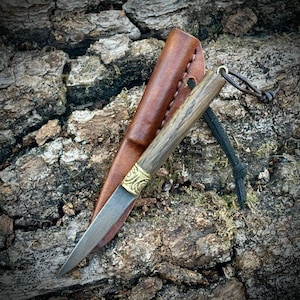 May include: A small, handcrafted knife with a wooden handle and a decorative brass bolster. The blade is silver, and the knife is paired with a brown leather sheath. A black leather cord is attached to the handle. The knife rests on a textured, brown surface.