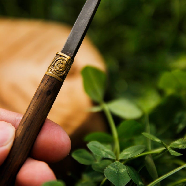 Puede incluir: Una hoja de metal oscuro unida a un mango cil&iacute;ndrico de madera. El mango presenta una veta de madera marr&oacute;n oscura texturizada y est&aacute; conectado a la hoja mediante una banda decorativa de metal dorado con un patr&oacute;n circular en relieve.