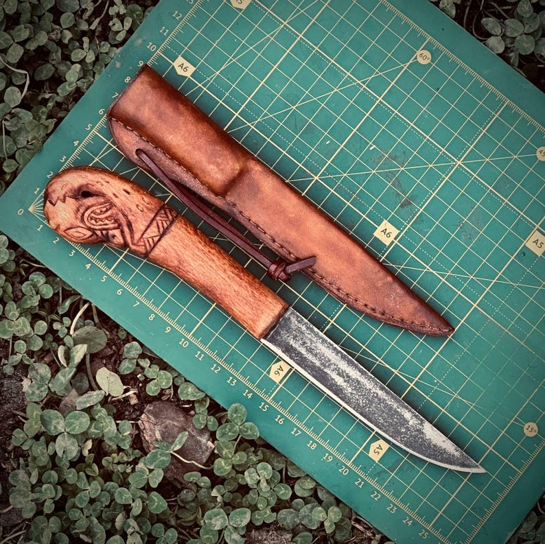 Op de afbeelding: Een handgemaakt mes met een houten handvat met gesneden details en een leren schede. Het lemmet heeft een rustieke, verouderde uitstraling. De schede is van bruin leer met een riem. Het mes ligt op een groene snijmat.