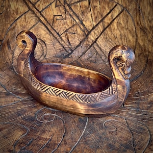 May include: A hand-carved wooden Viking ship bowl with detailed carvings. The bowl is brown with a dark interior and features intricate designs on the sides and ends. The ship has a curved prow and stern, and is set on a wooden surface.