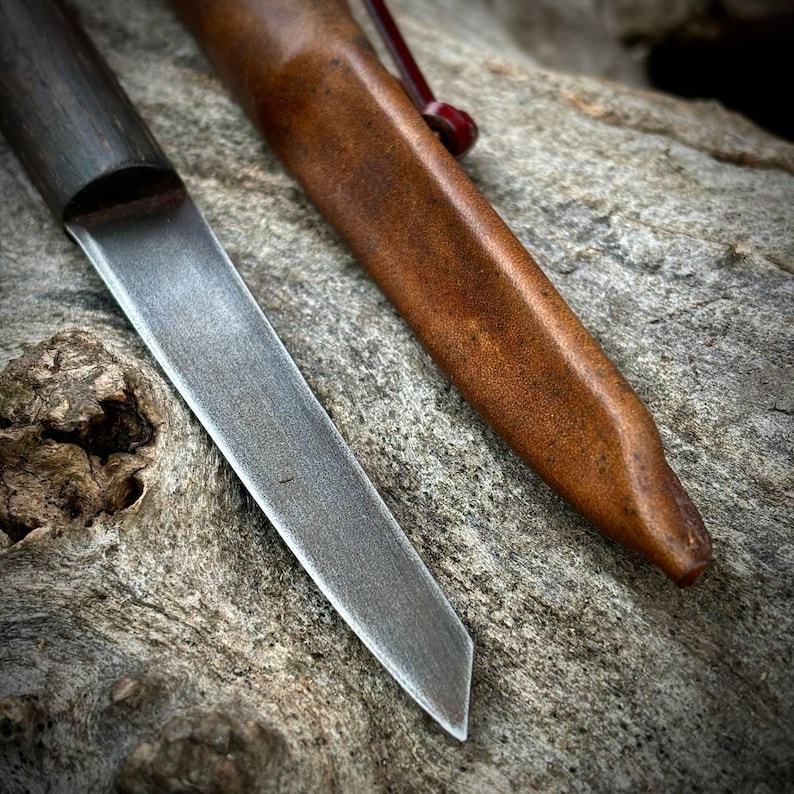 Puede incluir: Primer plano de un cuchillo afilado de plata con mango de madera y funda de cuero. El cuchillo est&aacute; sobre una superficie rugosa y texturizada.