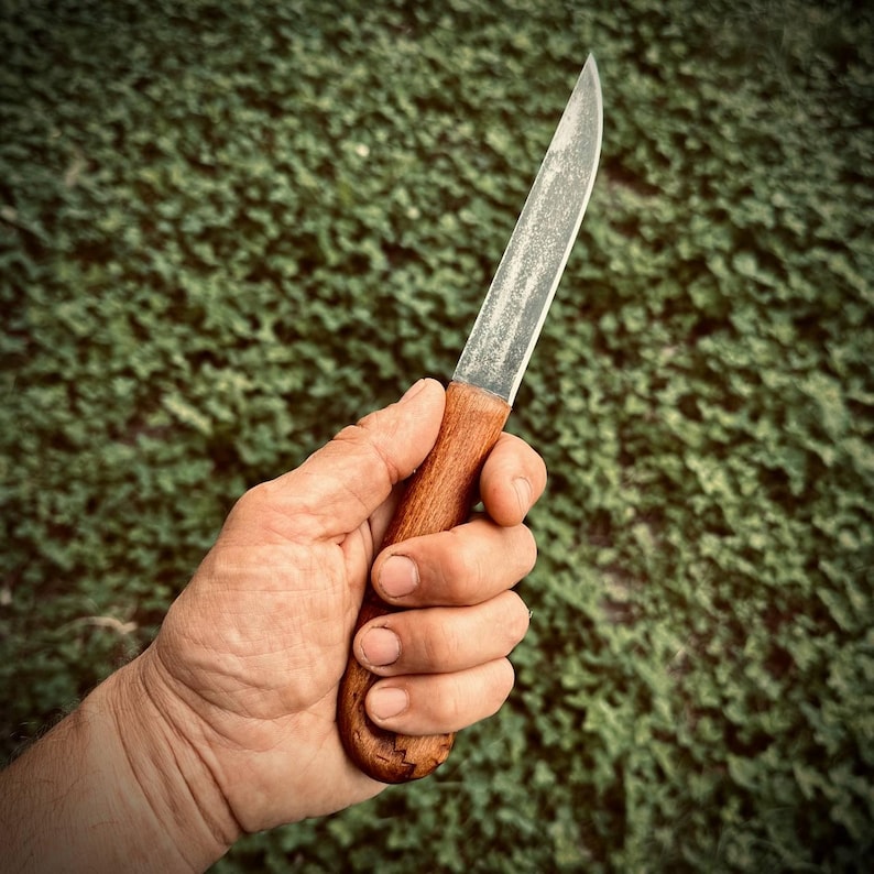 Op de afbeelding: Een mes met een houten handvat en een zilveren lemmet wordt in een hand gehouden. Het mes staat tegen een achtergrond van groen gebladerte.