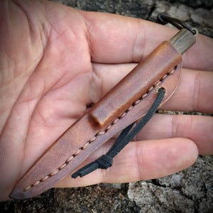 Puede incluir: Un cuchillo peque&ntilde;o dentro de una funda de cuero marr&oacute;n con costuras visibles a lo largo del borde. El mango del cuchillo es de madera oscura y cuenta con un cord&oacute;n de cuero negro atado alrededor de la funda para su sujeci&oacute;n.