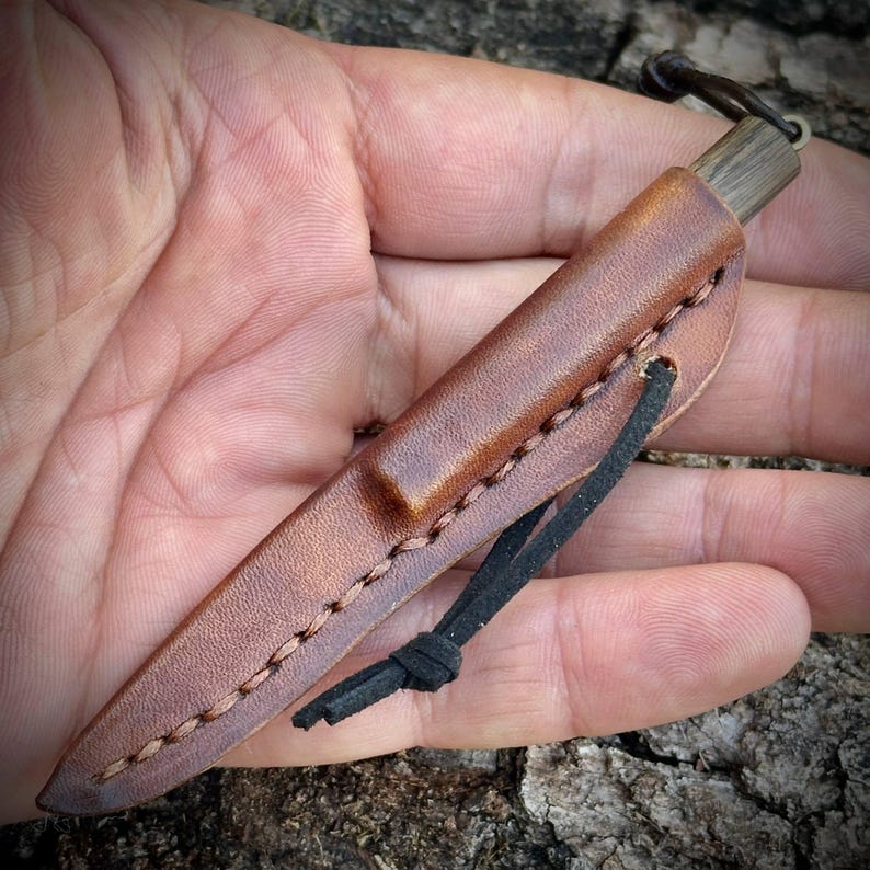 Puede incluir: Un cuchillo peque&ntilde;o dentro de una funda de cuero marr&oacute;n con costuras visibles a lo largo del borde. El mango del cuchillo es de madera oscura y cuenta con un cord&oacute;n de cuero negro atado alrededor de la funda para su sujeci&oacute;n.