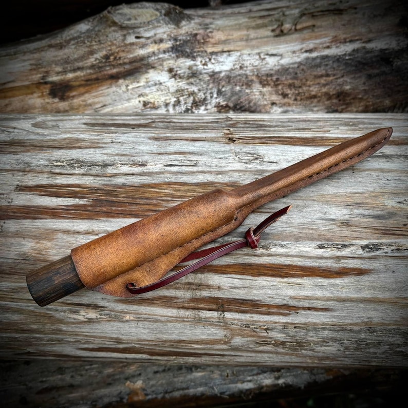Puede incluir: Una funda de cuero marr&oacute;n con un mango de madera y una correa de cuero roja. La funda est&aacute; dise&ntilde;ada para sostener un cuchillo u otro objeto afilado.