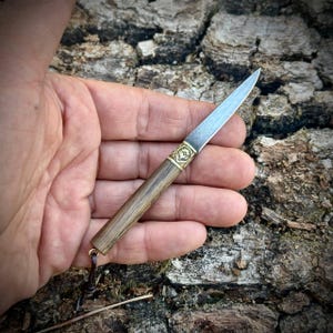 Puede incluir: Un cuchillo peque&ntilde;o de hoja fija con una hoja de acero puntiaguda, un refuerzo de lat&oacute;n con un patr&oacute;n floral grabado y un mango cil&iacute;ndrico de madera. Un cord&oacute;n de cuero oscuro est&aacute; unido al extremo del mango, que tiene una veta de madera visible.