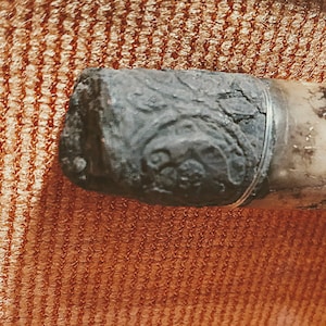 Puede incluir: Un objeto cil&iacute;ndrico que presenta una tapa de metal oscura, desgastada y texturizada con intrincados patrones en relieve de remolinos. La tapa est&aacute; unida a un cuerpo cil&iacute;ndrico m&aacute;s claro y liso, creando un contraste distintivo en color y textura.