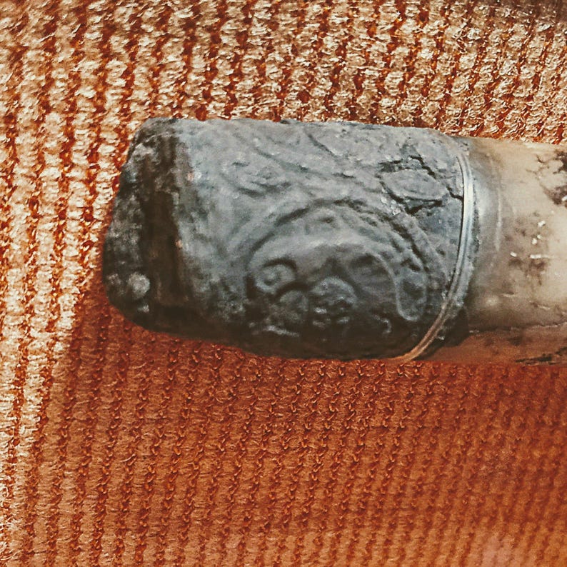 Puede incluir: Un objeto cil&iacute;ndrico que presenta una tapa de metal oscura, desgastada y texturizada con intrincados patrones en relieve de remolinos. La tapa est&aacute; unida a un cuerpo cil&iacute;ndrico m&aacute;s claro y liso, creando un contraste distintivo en color y textura.