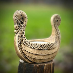 May include: A tan-colored, ceramic Viking ship planter with intricate carved details. The vessel features a dragon head at the prow and a curled stern. The sides are decorated with geometric patterns, and the planter sits on a wooden post against a blurred green background.