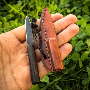 Puede incluir: Un cuchillo pequeño de hoja fija con mango de madera oscura y hoja metálica, acompañado de una funda de cuero marrón. Un cordón negro está unido al mango del cuchillo, lo que permite asegurarlo a la funda para su almacenamiento y transporte.