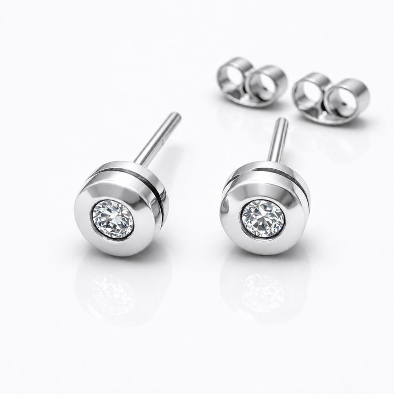 May include: A pair of silver-toned stud earrings featuring a circular bezel setting with a single clear, faceted round gemstone in the center. Each earring includes a post back and a separate matching silver-toned butterfly earring back.