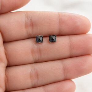 Puede incluir: Un par de pendientes de bot&oacute;n cuadrados peque&ntilde;os con piedras negras facetadas engastadas en monturas de metal plateado con un borde texturizado y perlado. Los pendientes tienen una forma geom&eacute;trica y un acabado pulido.