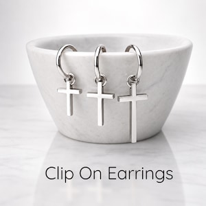 Puede incluir: Tres pendientes de clip de metal plateado, cada uno con un pequeño dije de cruz colgando de un aro circular. El texto "Clip On Earrings" está impreso en negro en la parte inferior de la imagen, que muestra estos artículos de joyería.
