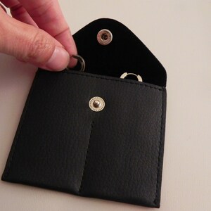 K&ouml;nnte beinhalten: Ein schwarzes Lederetui mit zwei F&auml;chern zur Aufbewahrung von kleinen Gegenst&auml;nden wie Schmuck. Das Etui hat einen Druckknopfverschluss und zwei silberne Metalldruckkn&ouml;pfe.