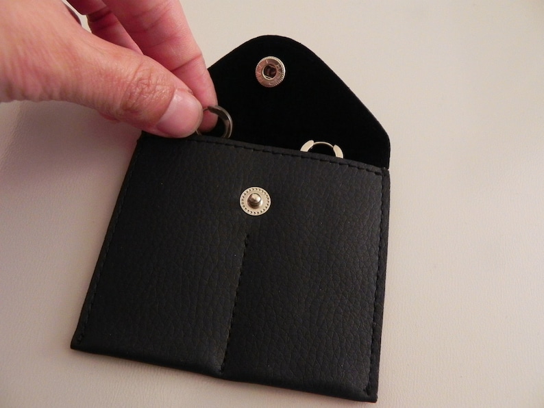 K&ouml;nnte beinhalten: Ein schwarzes Lederetui mit zwei F&auml;chern zur Aufbewahrung von kleinen Gegenst&auml;nden wie Schmuck. Das Etui hat einen Druckknopfverschluss und zwei silberne Metalldruckkn&ouml;pfe.