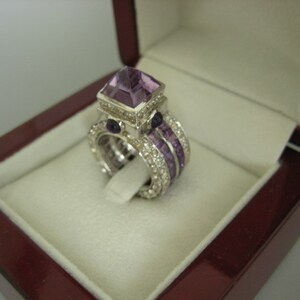 K&ouml;nnte beinhalten: Ein silberner Ring mit einem gro&szlig;en, quadratischen Amethyst-Edelstein, der von kleineren Amethyst-Steinen und Diamanten umgeben ist. Der Ring befindet sich in einer burgunderfarbenen Samt-Ringbox.