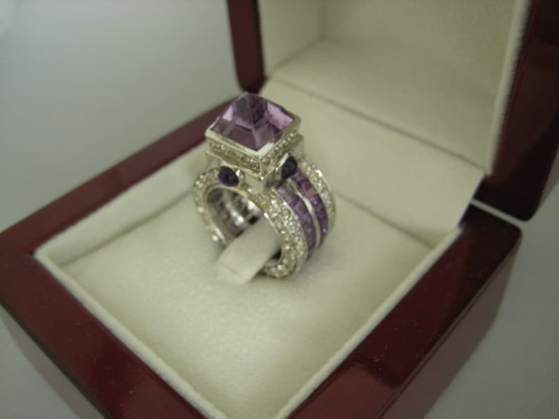 K&ouml;nnte beinhalten: Ein silberner Ring mit einem gro&szlig;en, quadratischen Amethyst-Edelstein, der von kleineren Amethyst-Steinen und Diamanten umgeben ist. Der Ring befindet sich in einer burgunderfarbenen Samt-Ringbox.