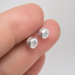 May include: A pair of small, round, silver-toned stud earrings, each featuring a single clear, faceted stone set securely within a smooth, polished metal bezel. The earrings have a clean, circular design with a reflective surface finish.