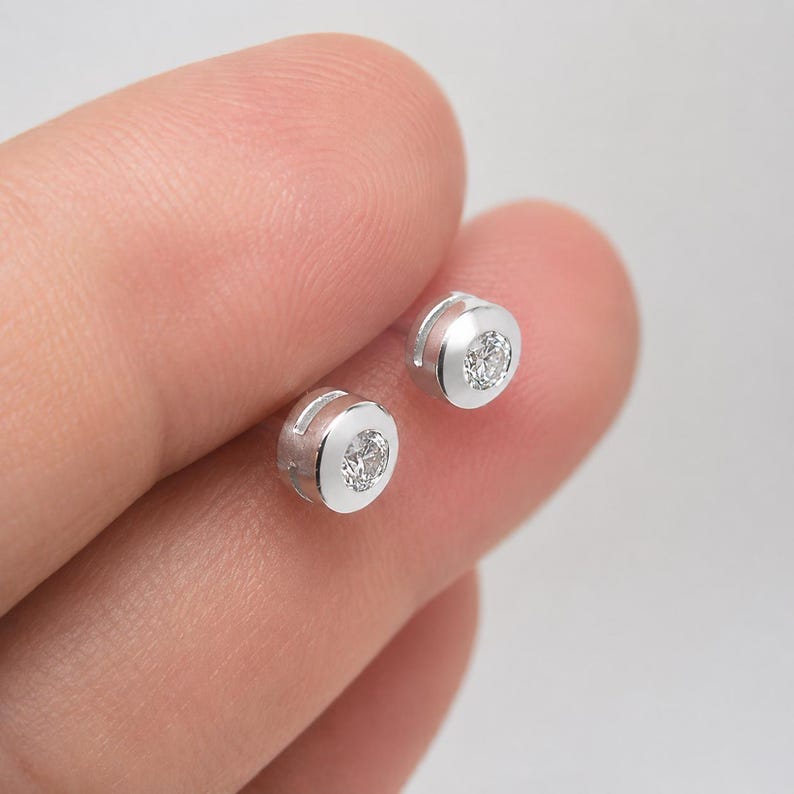 May include: A pair of small, round, silver-toned stud earrings, each featuring a single clear, faceted stone set securely within a smooth, polished metal bezel. The earrings have a clean, circular design with a reflective surface finish.