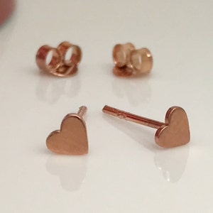 May include: A pair of rose gold heart-shaped stud earrings. The earrings are small and delicate, and they have a simple, elegant design.