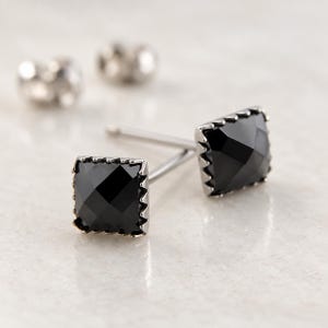 Puede incluir: Un par de pendientes de bot&oacute;n cuadrados con piedras negras facetadas engastadas en un marco met&aacute;lico con borde dentado. Los pendientes incluyen postes met&aacute;licos finos y cierres met&aacute;licos a juego con un acabado pulido.