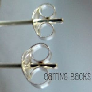 Może przedstawiać: Dwa srebrne zapięcia do kolczyków, zwane również sztyftami do kolczyków, pokazane są na zbliżeniu. Zapięcia do kolczyków są wykonane z metalu i mają gładkie, błyszczące wykończenie.