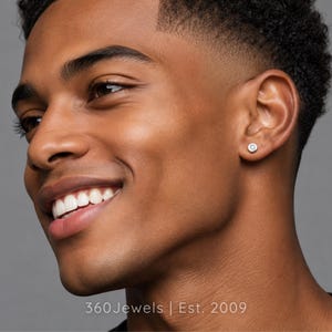 May include: A small, circular silver-toned stud earring featuring a single clear, brilliant-cut gemstone set in the center. The earring is worn on the earlobe. The text 360Jewels | Est. 2009 is visible at the bottom of the image.