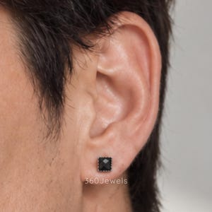 Puede incluir: Un pendiente de bot&oacute;n negro de forma cuadrada con una superficie facetada y un borde met&aacute;lico, colocado en el l&oacute;bulo de la oreja. El texto 360Jewels es visible en letras blancas debajo del pendiente. La piedra oscura y pulida est&aacute; engastada.
