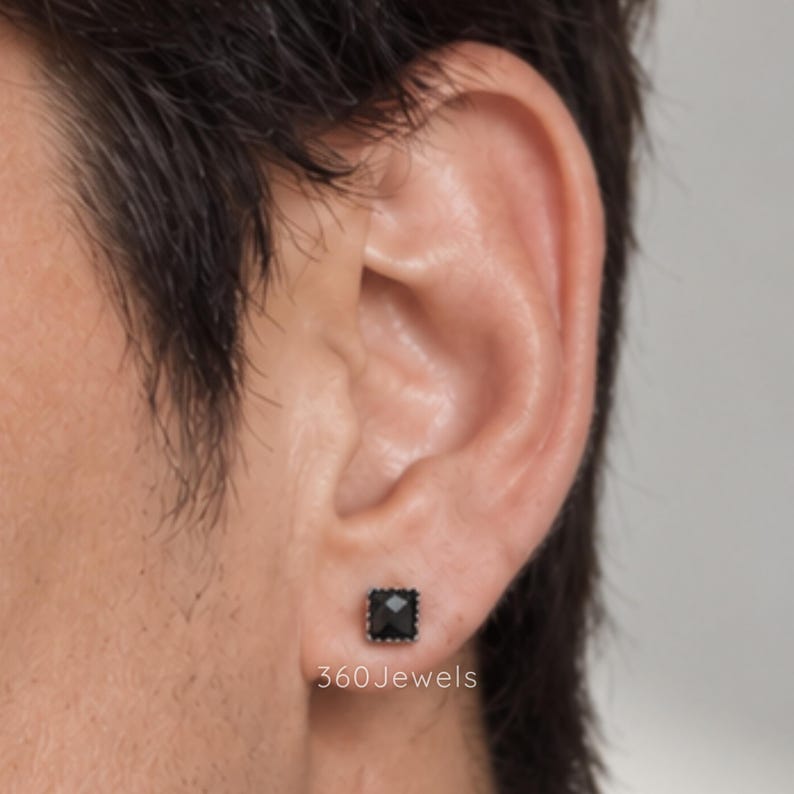 Puede incluir: Un pendiente de bot&oacute;n negro de forma cuadrada con una superficie facetada y un borde met&aacute;lico, colocado en el l&oacute;bulo de la oreja. El texto 360Jewels es visible en letras blancas debajo del pendiente. La piedra oscura y pulida est&aacute; engastada.