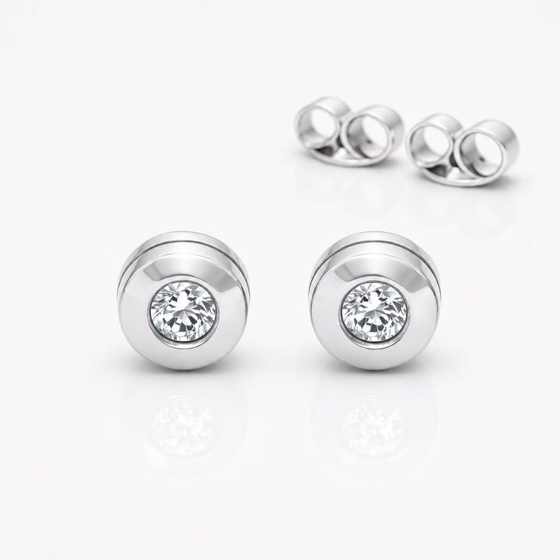 May include: A pair of silver-toned stud earrings featuring a circular bezel setting that holds a single clear, round-cut gemstone in the center. The earrings include matching silver-toned butterfly-style earring backs positioned behind them.