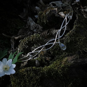 Puede incluir: Una tiara plateada con un diseño intrincado y fluido, adornada con pequeños colgantes de cristal transparente. La tiara descansa sobre un lecho de musgo, junto a una flor blanca con centro amarillo y hojas verdes.