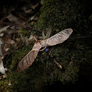 May include: A copper-toned pendant necklace featuring a stylized maple seed design. The pendant has two wing-shaped elements with detailed veining, suspended from a delicate chain. A small, beaded charm hangs below the seed. The necklace rests on a bed of green moss.