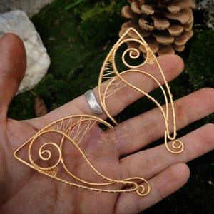 May include: A pair of gold-colored wire elf ear cuffs. The cuffs are held in a hand, and feature intricate wirework with spiral and linear designs. The background includes moss, a pinecone, and a rock.