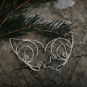 May include: A silver wire mask with a delicate, intricate design resembling a butterfly. The mask is set against a textured, gray background.