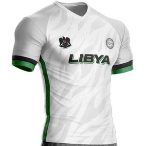 Può includere: Maglia da calcio bianca e grigia con dettagli verdi e neri. La maglia presenta la scritta "LIBYA" in lettere verdi e nere, uno stemma e un emblema circolare. Le maniche hanno bande verdi e nere.