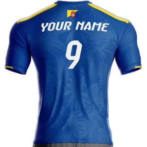 K&ouml;nnte beinhalten: Blaues Sporttrikot mit gelben Akzenten an Schultern und &Auml;rmeln. Auf der R&uuml;ckseite befindet sich die wei&szlig;e Nummer 9 und der Text "YOUR NAME". Ein Wappen ist in der N&auml;he des Kragens zu sehen.