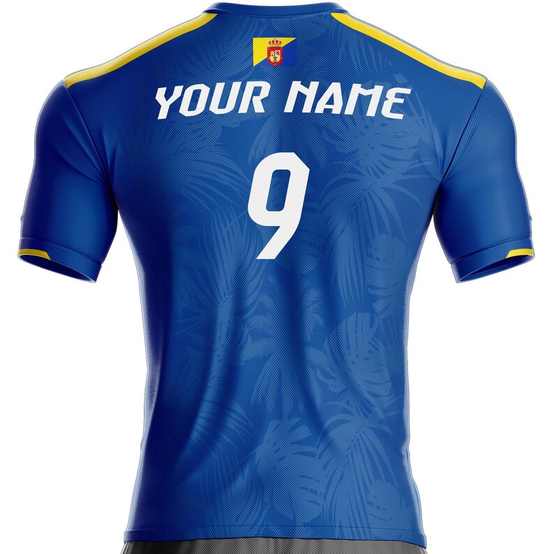 K&ouml;nnte beinhalten: Blaues Sporttrikot mit gelben Akzenten an Schultern und &Auml;rmeln. Auf der R&uuml;ckseite befindet sich die wei&szlig;e Nummer 9 und der Text "YOUR NAME". Ein Wappen ist in der N&auml;he des Kragens zu sehen.
