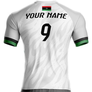 Può includere: Maglia da calcio bianca con dettagli neri e verdi. La maglia presenta il numero 9 in nero, la scritta "YOUR NAME" in nero e una bandiera libica sulla parte superiore della schiena. Le maniche hanno finiture verdi e nere.