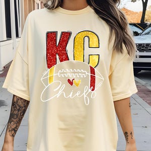 May include: Cream-colored t-shirt featuring the text "KC Chiefs" in red and yellow. The design includes a football graphic with red and yellow hearts. The shirt has short sleeves and a relaxed fit.